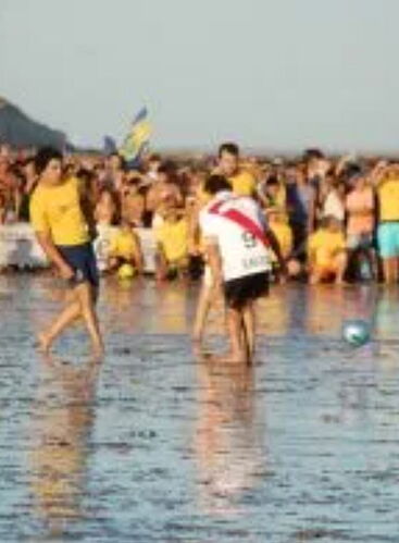 La imagen muestra una impresionante vista aérea de una playa con palmeras y una <tag>person</tag> caminando solo a lo largo de la orilla, con el sol brillo en el fondo y unas pocas <tag>boat</tag> ancladas en el mar. (Subtitulado por IA)