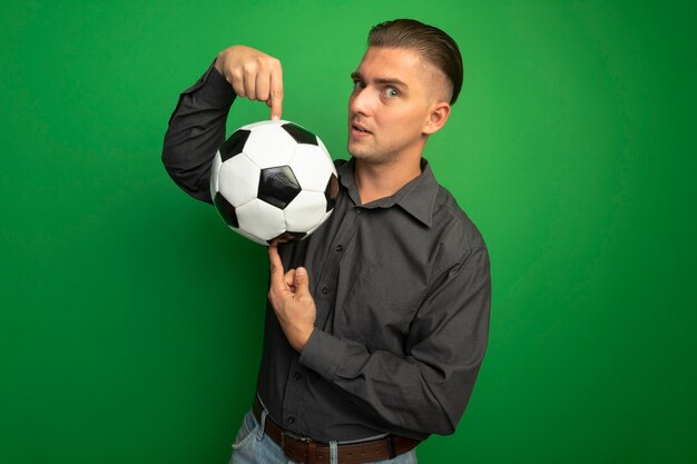 hombre-guapo-joven-camisa-gris-que-muestra-balon-futbol-apuntando-dedo-indice-sonriendo-confiado-pie-sobre-pared-verde_141793-53484